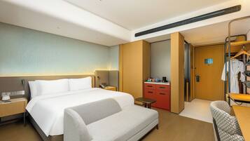 A hotel room with a large bed, a sofa, a desk, a small table, a mirror, a wardrobe, and a bathroom door.