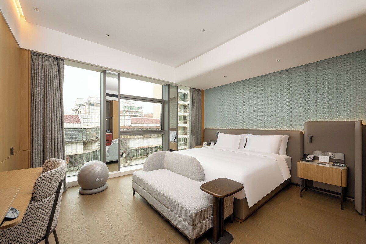 A modern hotel room with a large bed, a sofa, a small table, and a view of the city through large windows.