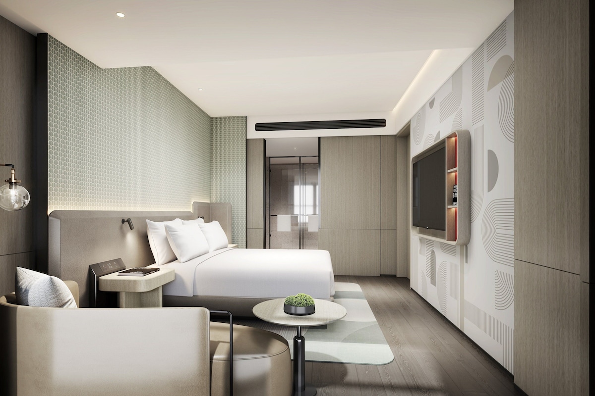A modern hotel room with a bed, a desk, a chair, a TV, and a small table with a plant.