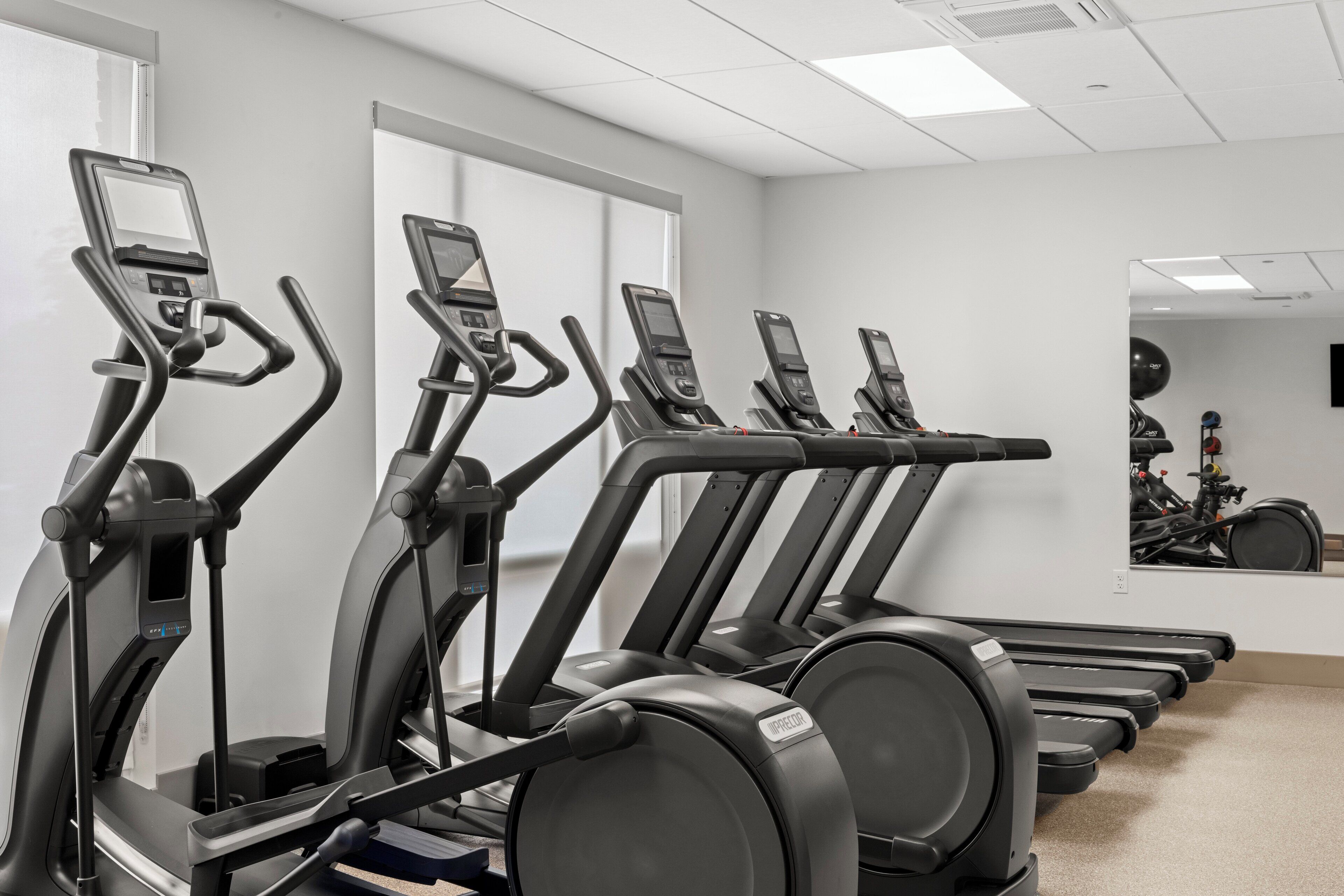 Eine Reihe von Steh- oder Heimtrainern in einem Fitnessstudio, wobei sich die Geräte in einem großen Spiegel spiegeln.