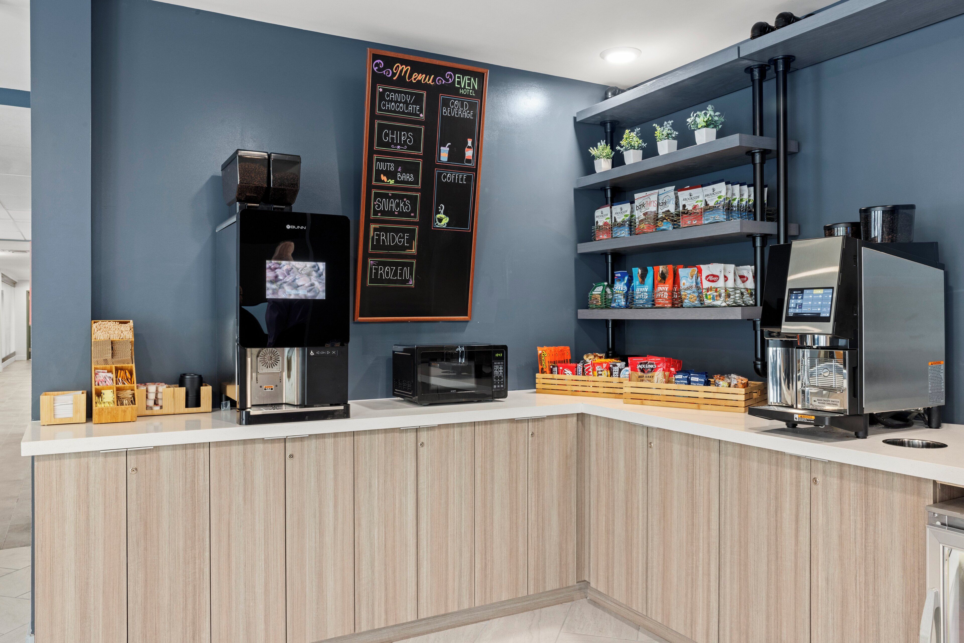 A modern kitchen area with a coffee machine, microwave, and a chalkboard menu listing items like snacks, cold beverages, and coffee.
