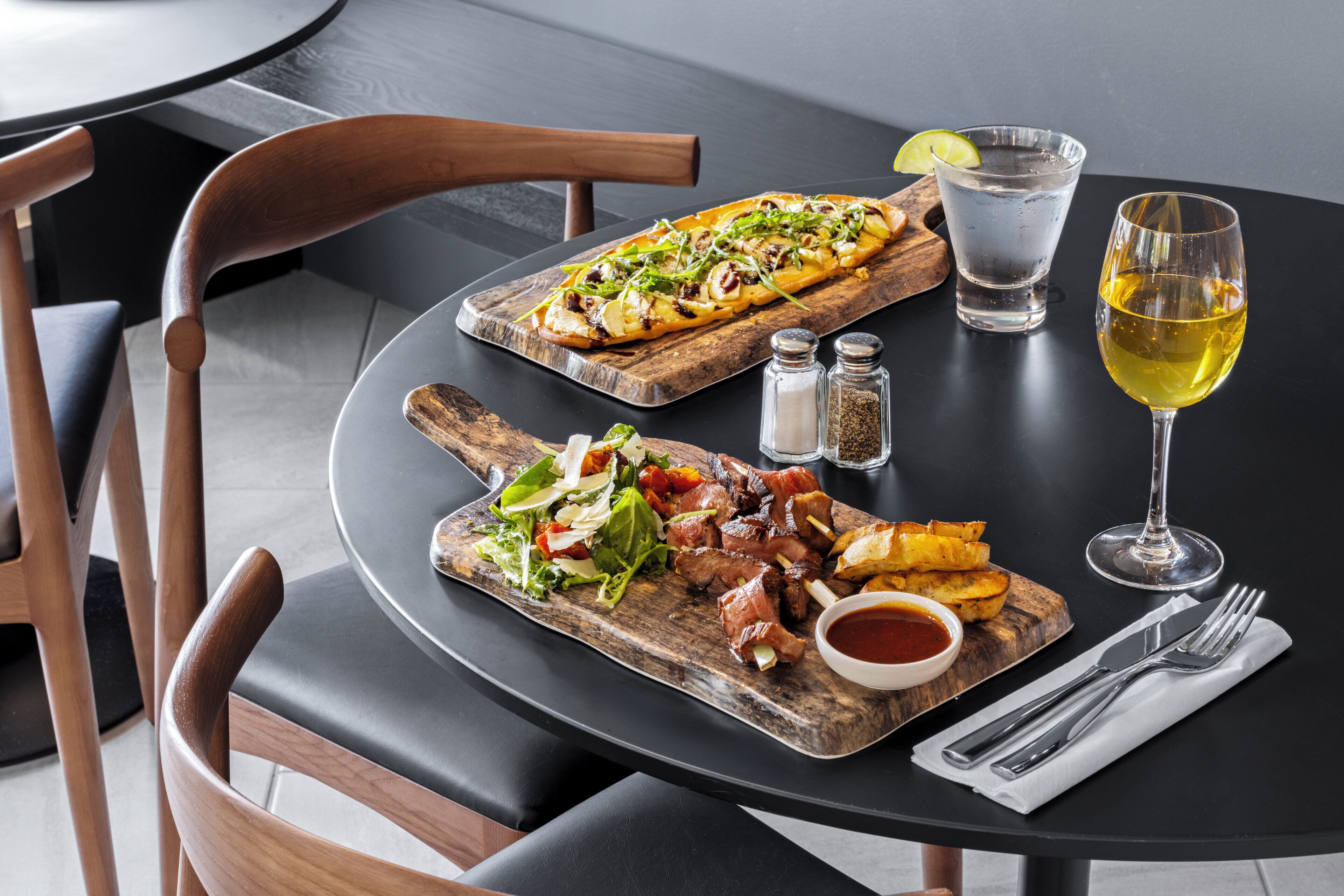 A dining table with a pizza, a salad, and a steak dish. There are salt and pepper shakers, a glass of water, and a glass of wine on the table.