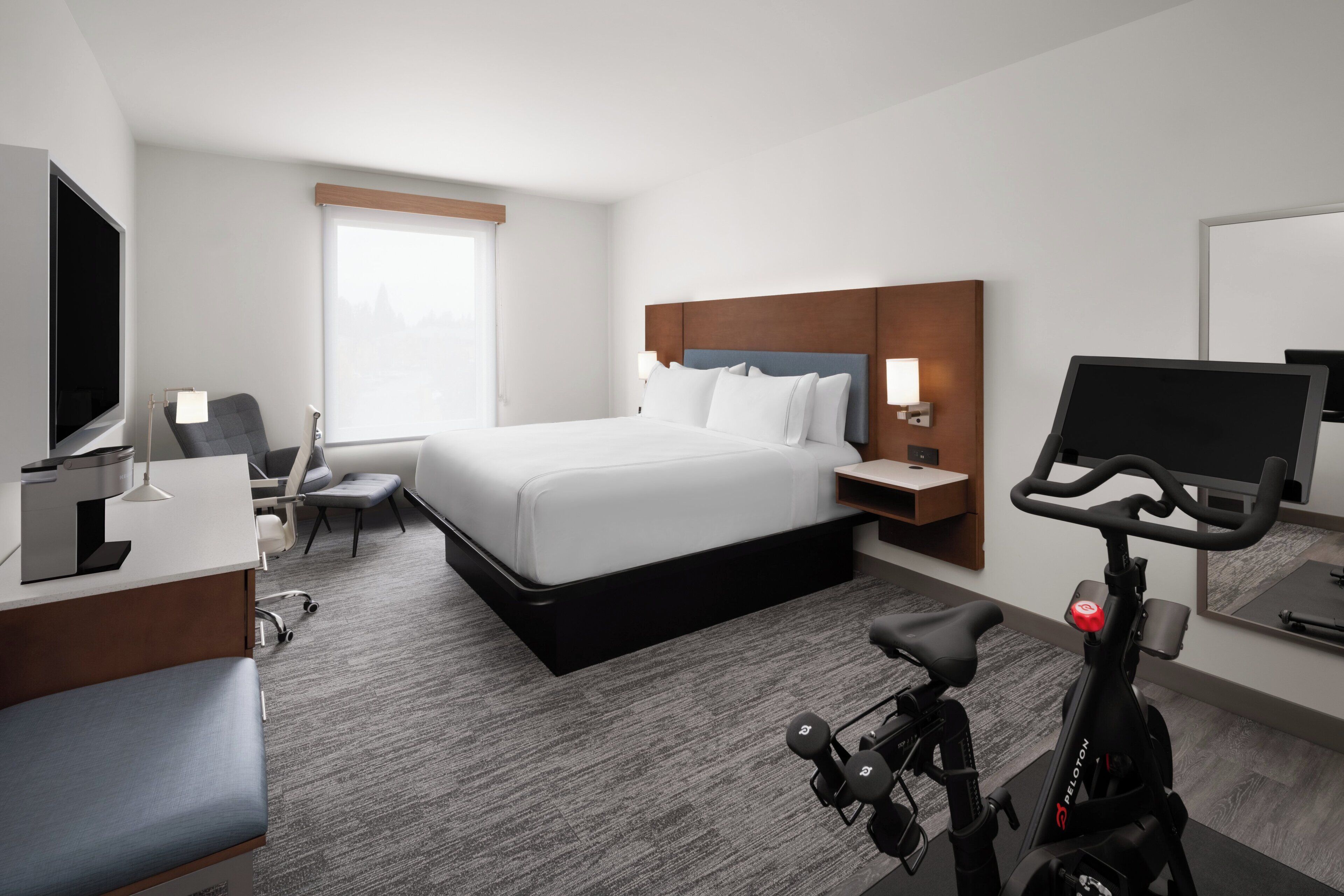 A hotel room with a bed, desk, chair, TV, exercise bike, and a window.