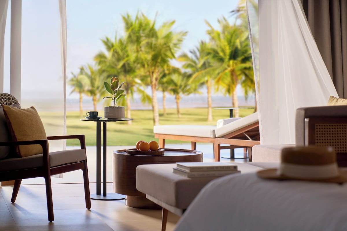 Una habitación de hotel con cama, una silla, una mesita con una planta y vistas a las palmeras y al océano.