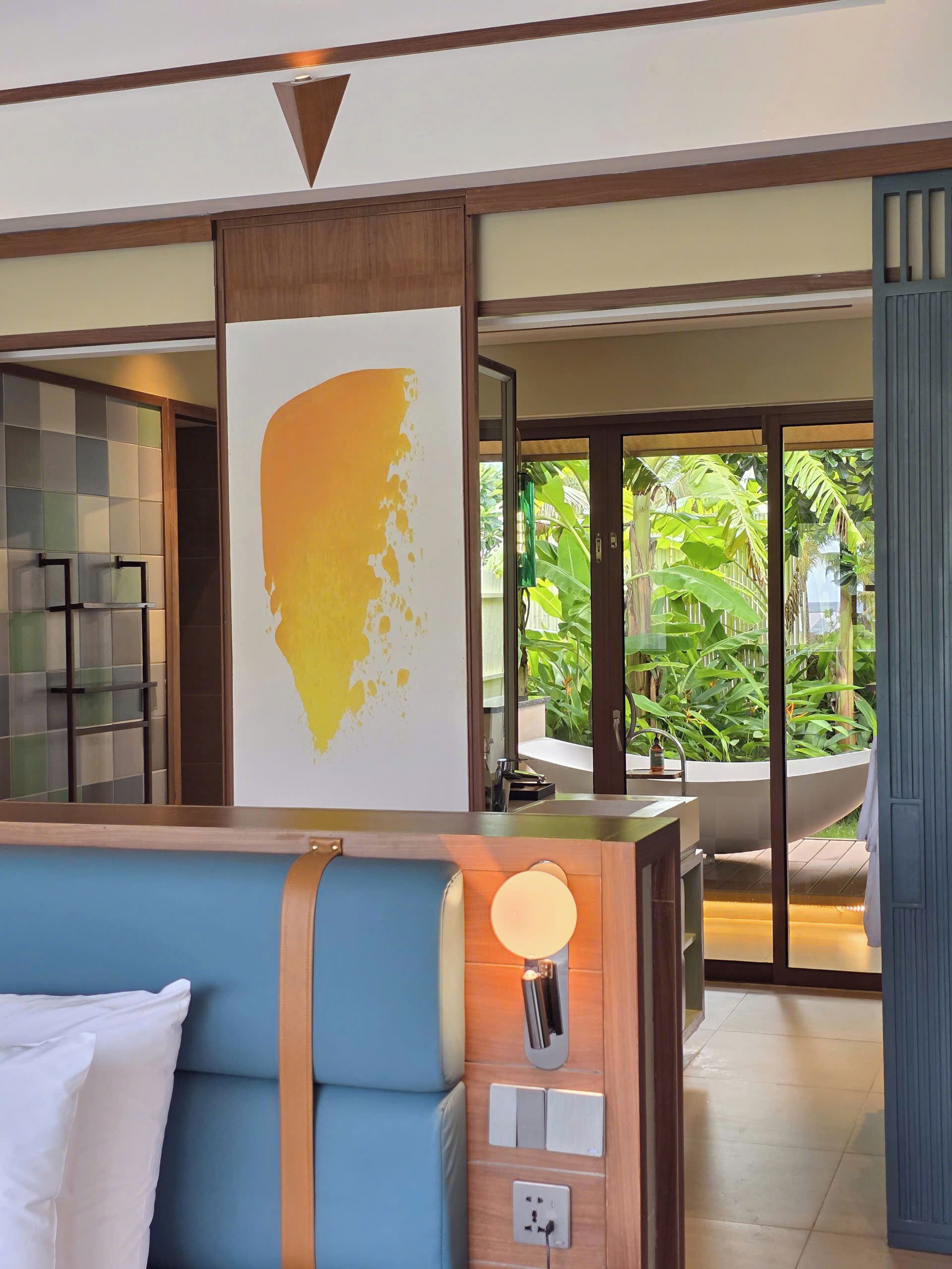 A hotel room with a large bed, a bedside table with a lamp, a view of a garden through glass doors, and a wall with a large yellow artwork.