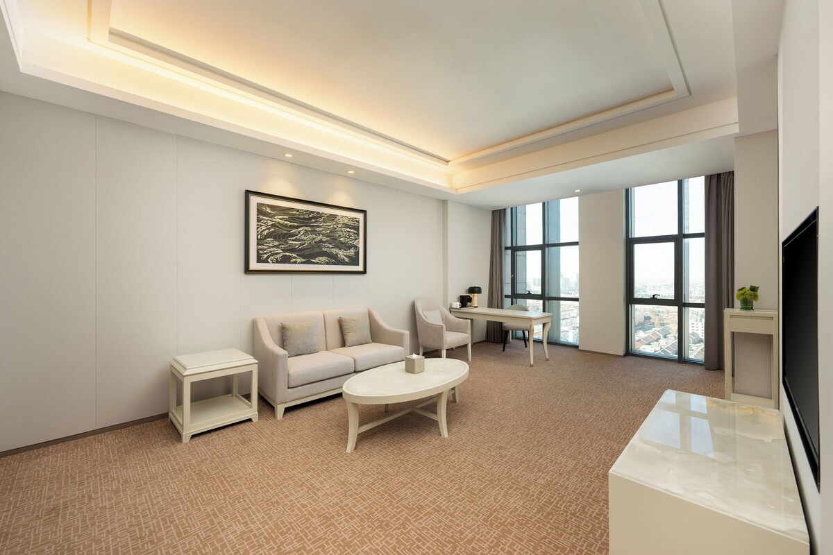 Une chambre d’hôtel moderne dotée d’un canapé, d’une table basse, d’une télévision et de grandes fenêtres offrant une vue sur la ville.