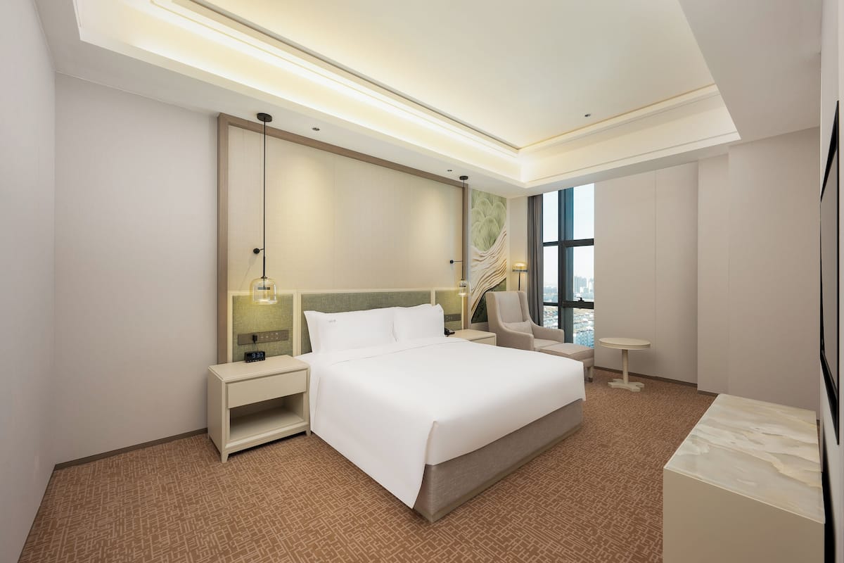 A modern hotel room with a large bed, a sitting area with a chair and a small table, a large mirror, and a city view through the window.