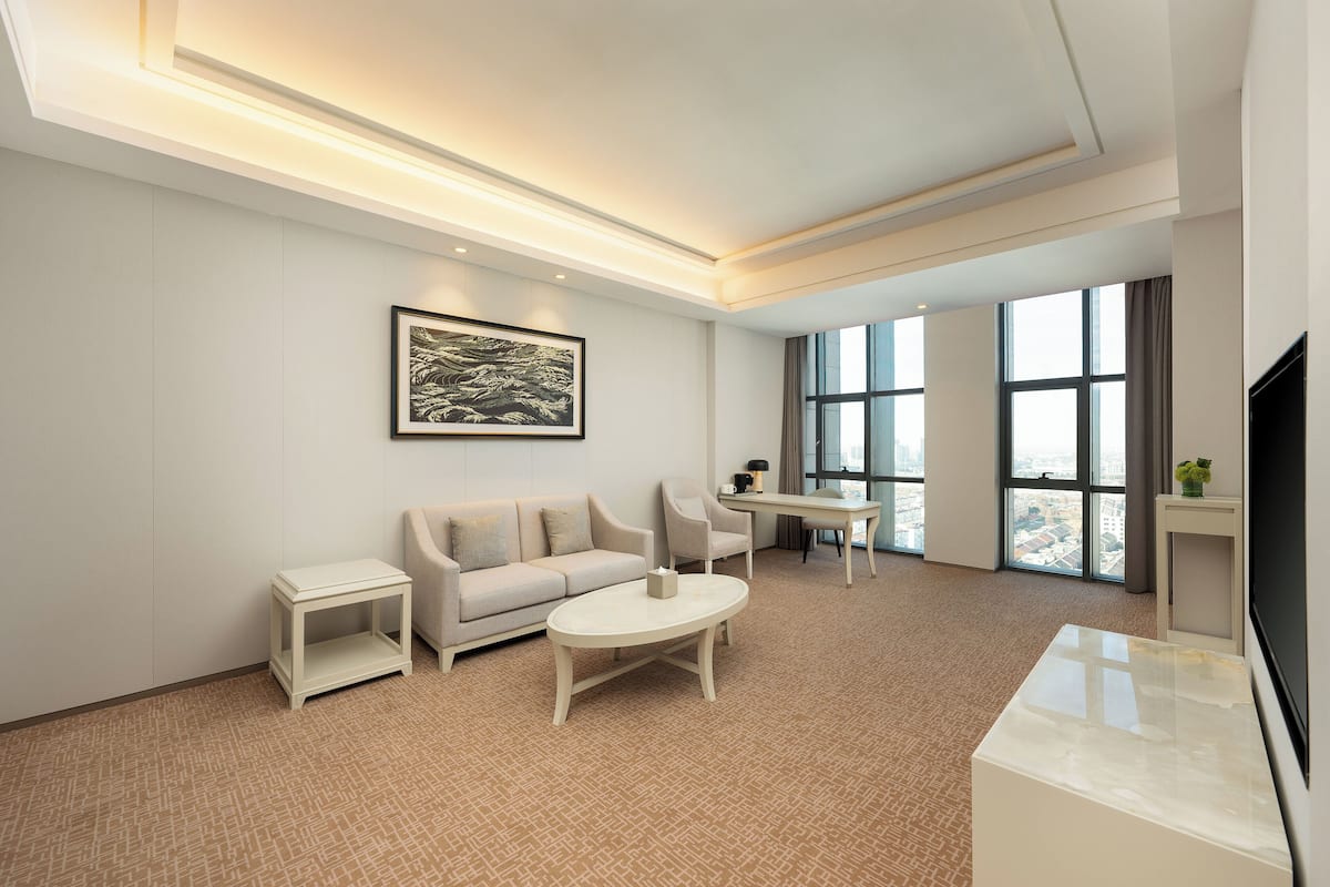 A modern hotel room with a sofa, a coffee table, a TV, and large windows offering a city view.