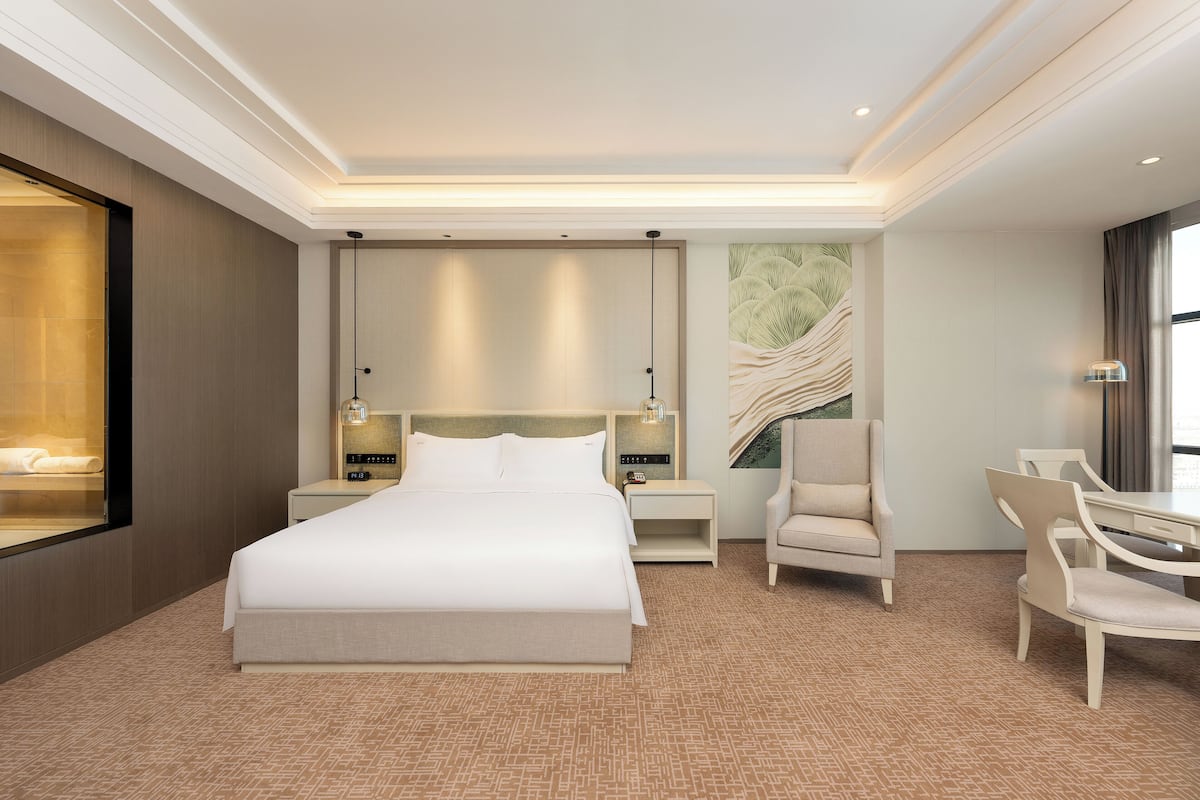 A hotel room with a large bed, bedside tables, a chair, a desk, and a large window.
