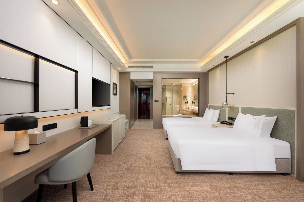 Une chambre d’hôtel équipée d’un bureau, de deux lits, d’une télévision et d’une salle de bain visible à travers une porte en verre.