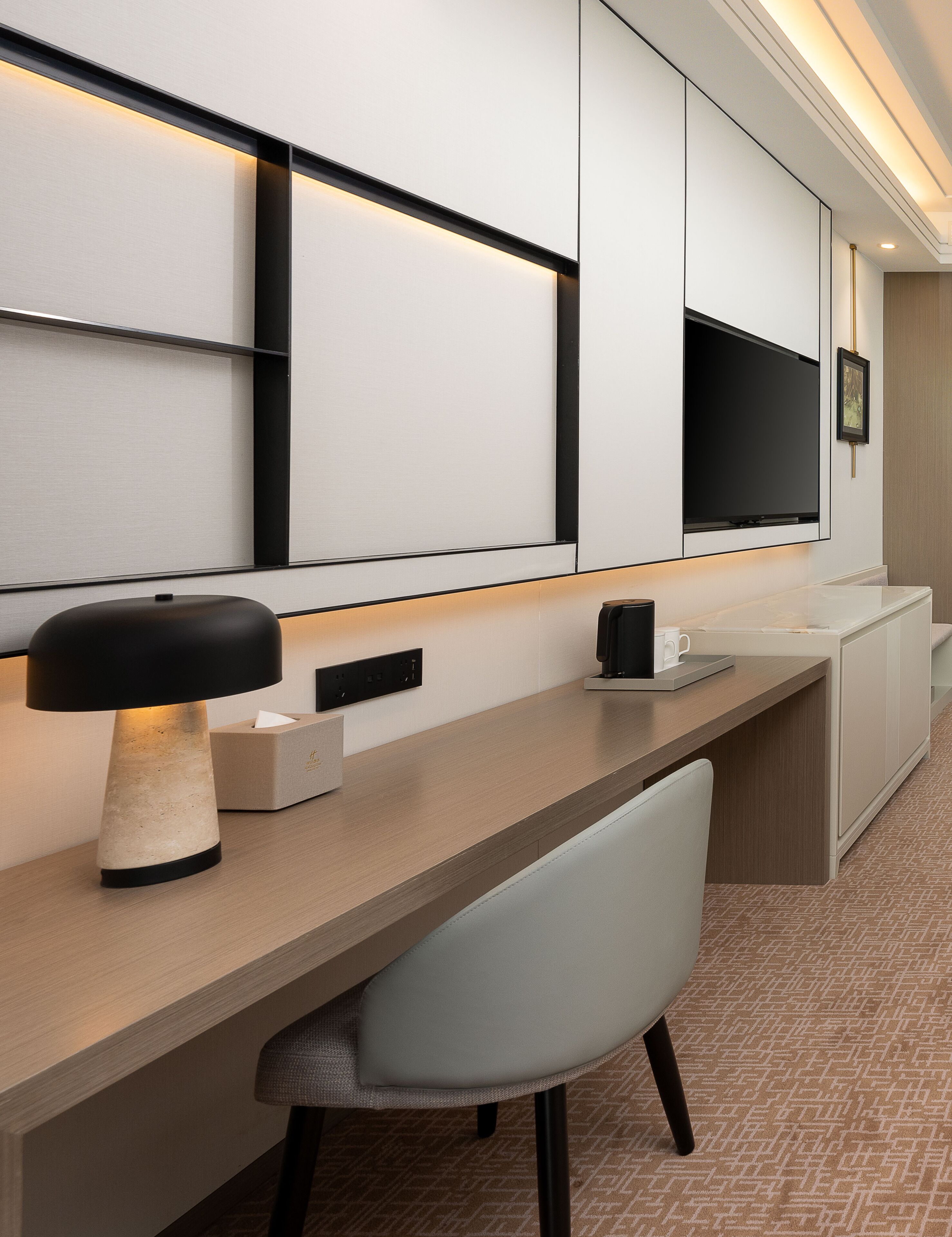 Habitación de hotel moderna con un escritorio grande, una silla, un televisor de pantalla plana montado en la pared y una mesita de noche con una lámpara y una caja de pañuelos.