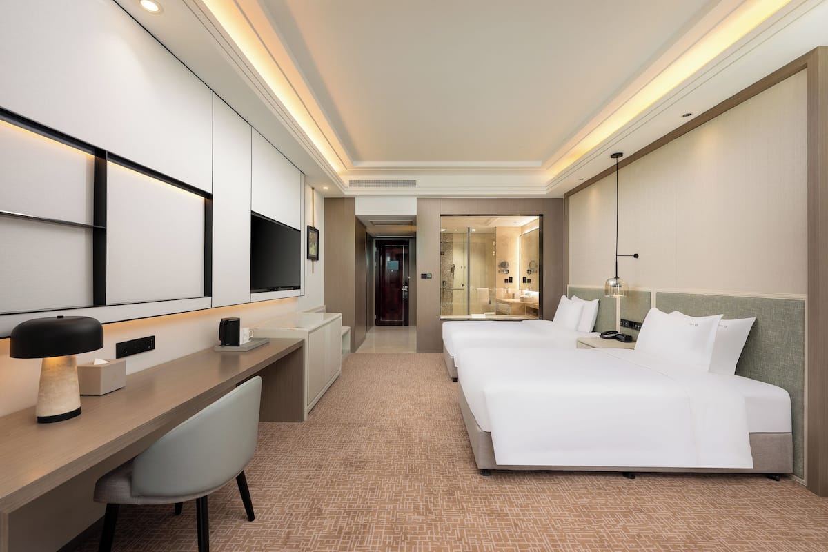 A hotel room with a desk, two beds, a TV, and a bathroom visible through a glass door.