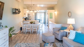Un salon moderne avec un canapé gris, une table à manger blanche avec des chaises, une table basse avec un panier, et une cuisine en arrière-plan.