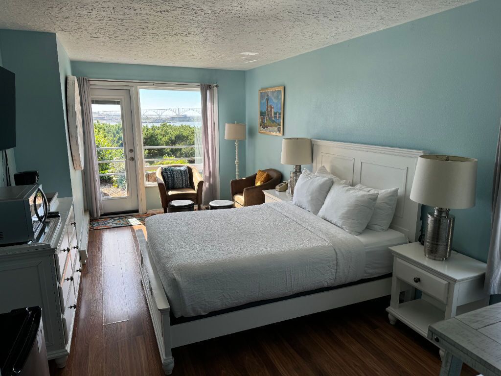 A bedroom with a large bed, a nightstand, a desk, and a chair. There is a window with a view of greenery and a building outside.