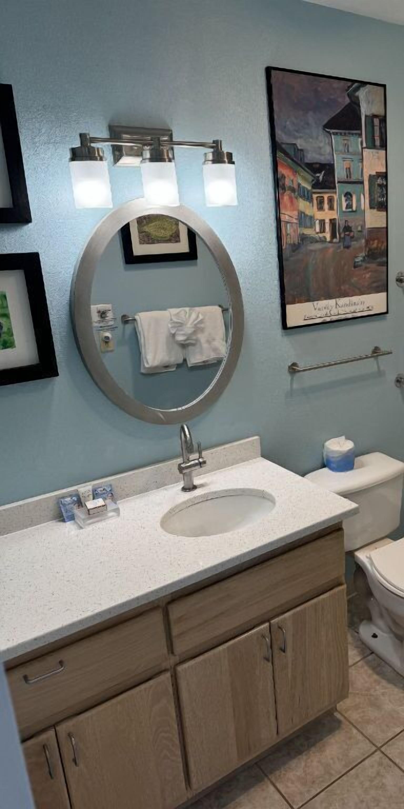 Een badkamer met een ronde spiegel, een wastafel, een toilet en een schilderij in een lijst aan de muur.