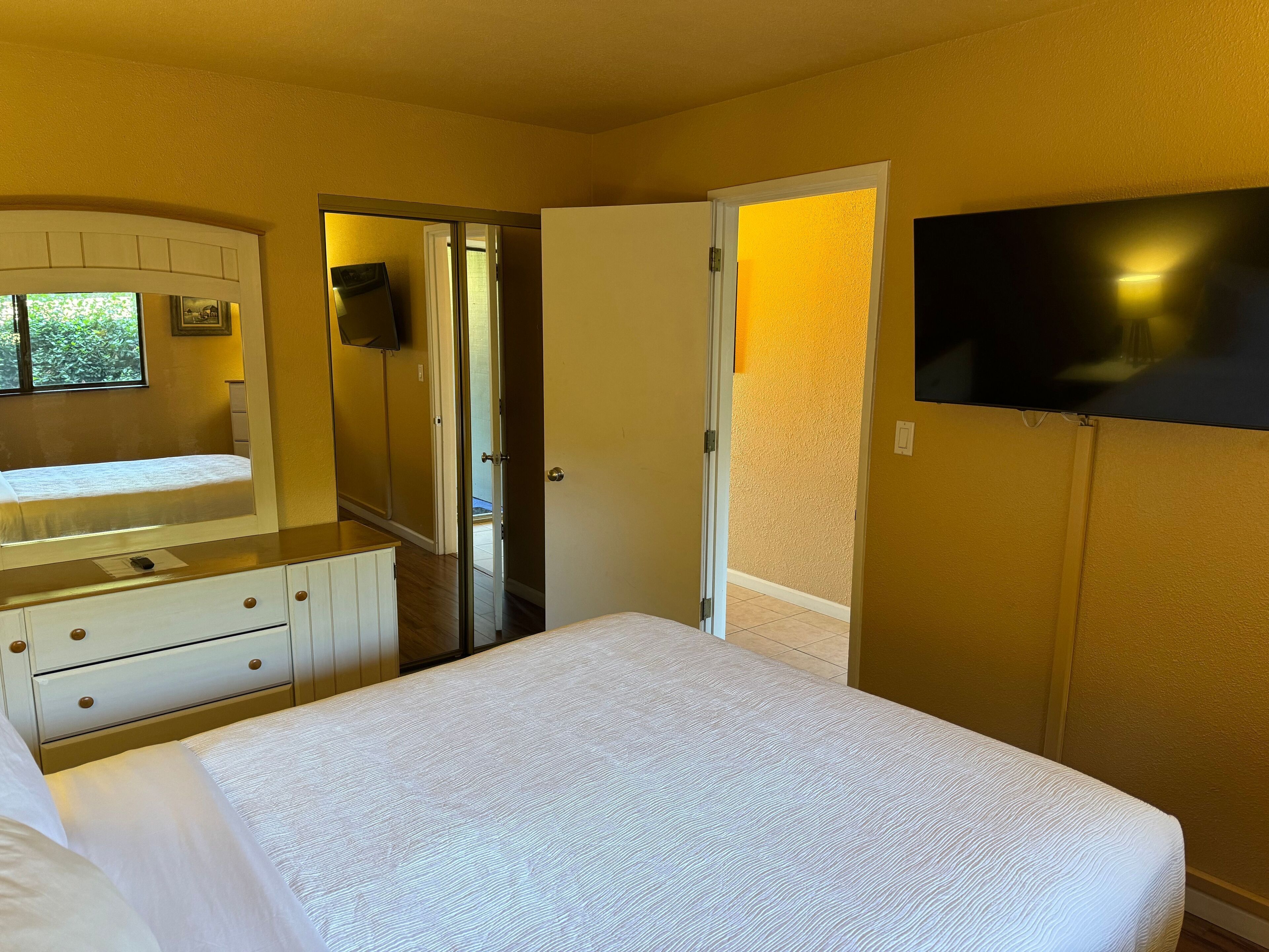 Une chambre à coucher comprenant un lit, une commode, un téléviseur fixé au mur et une porte donnant sur une autre pièce.
