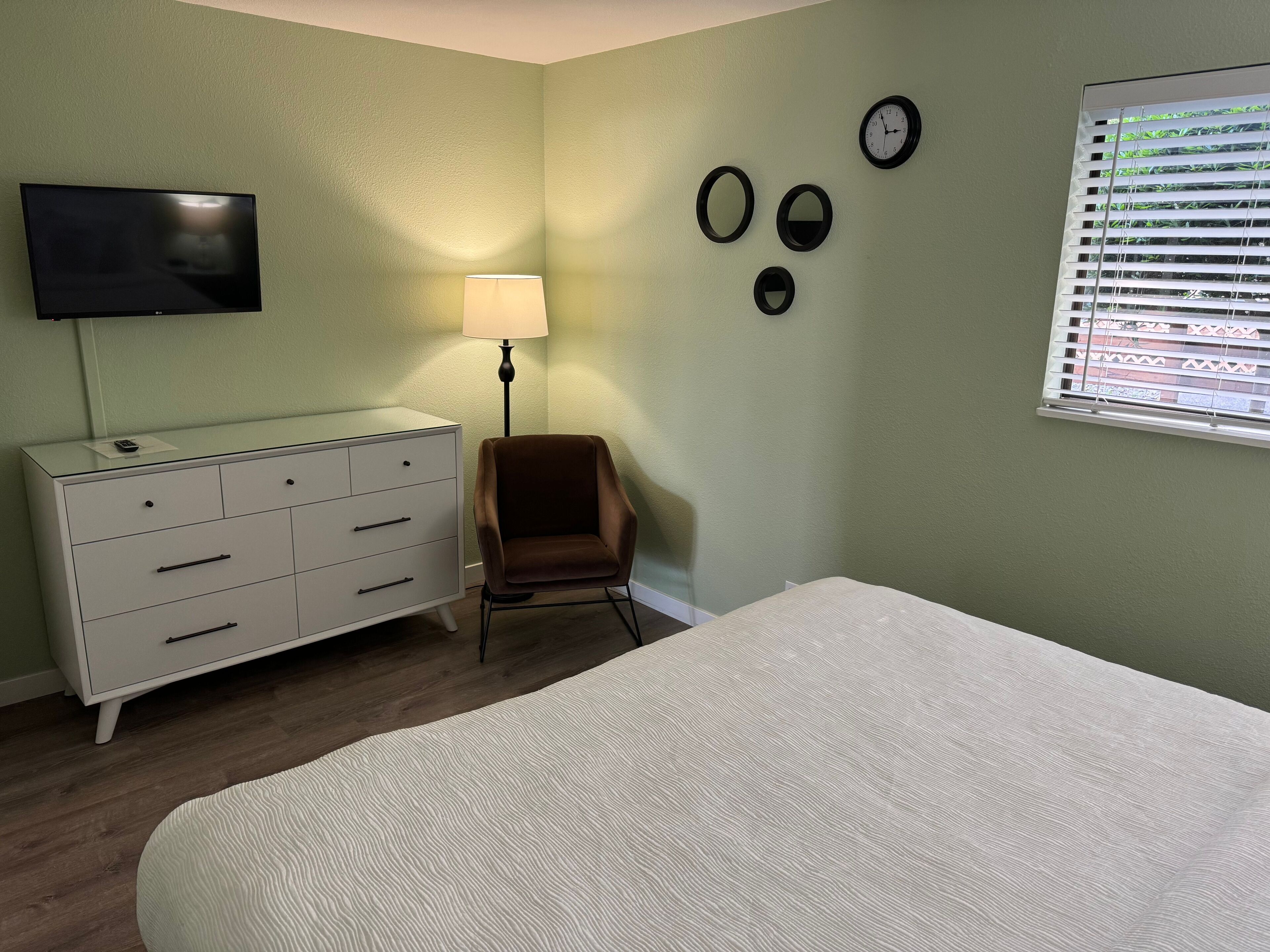 Un dormitorio con cama, cómoda, silla, lámpara y un reloj de pared.