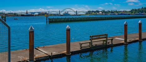 A waterfront dock with a bench, wooden bollards, and a bridge in the background.