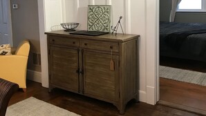 Una habitació amb una paret fosca, un armari de fusta amb objectes decoratius, una cadira groga i un llit amb llençols foscos.
