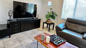 A modern living room with a flat-screen TV on a black entertainment unit, a leather sofa, a wooden coffee table with a board game, a potted plant, and a neon sign on the wall. - BALLOON FESTIVAL experience ABQ in style newly renovated 2BD/1BA apartment (Albuquerque)