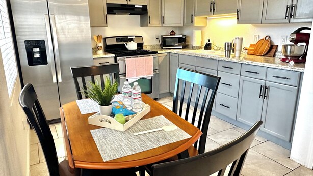 A modern kitchen with grey cabinets, a stainless steel refrigerator, a microwave, and a stove. There is a dining table with chairs, a potted plant, and a tray with food and drinks. - BALLOON FESTIVAL experience ABQ in style newly renovated 2BD/1BA apartment (Albuquerque)