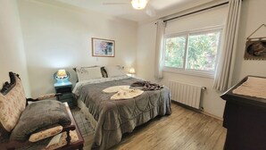 Quarto com cama, cadeira, mesa de cabeceira com abajur, radiador e janela com cortinas.