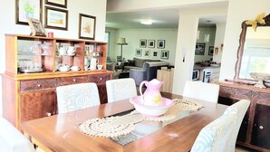 Uma área de refeições com mesa de madeira, cadeiras e um jarro decorativo. Há um armário com copos e um abajur ao fundo.