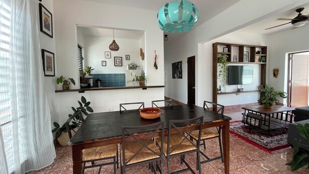 Un comedor con una mesa de madera oscura y sillas tejidas, una lámpara colgante color turquesa, y una cocina al fondo.