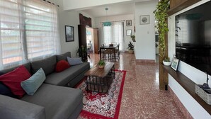 Amplio salón con un sofá gris, cojines rojos y azules, una mesa de centro de madera y una alfombra roja estampada. Hay un rincón comedor con una mesa y sillas, y un televisor de pantalla plana grande montado en la pared.
