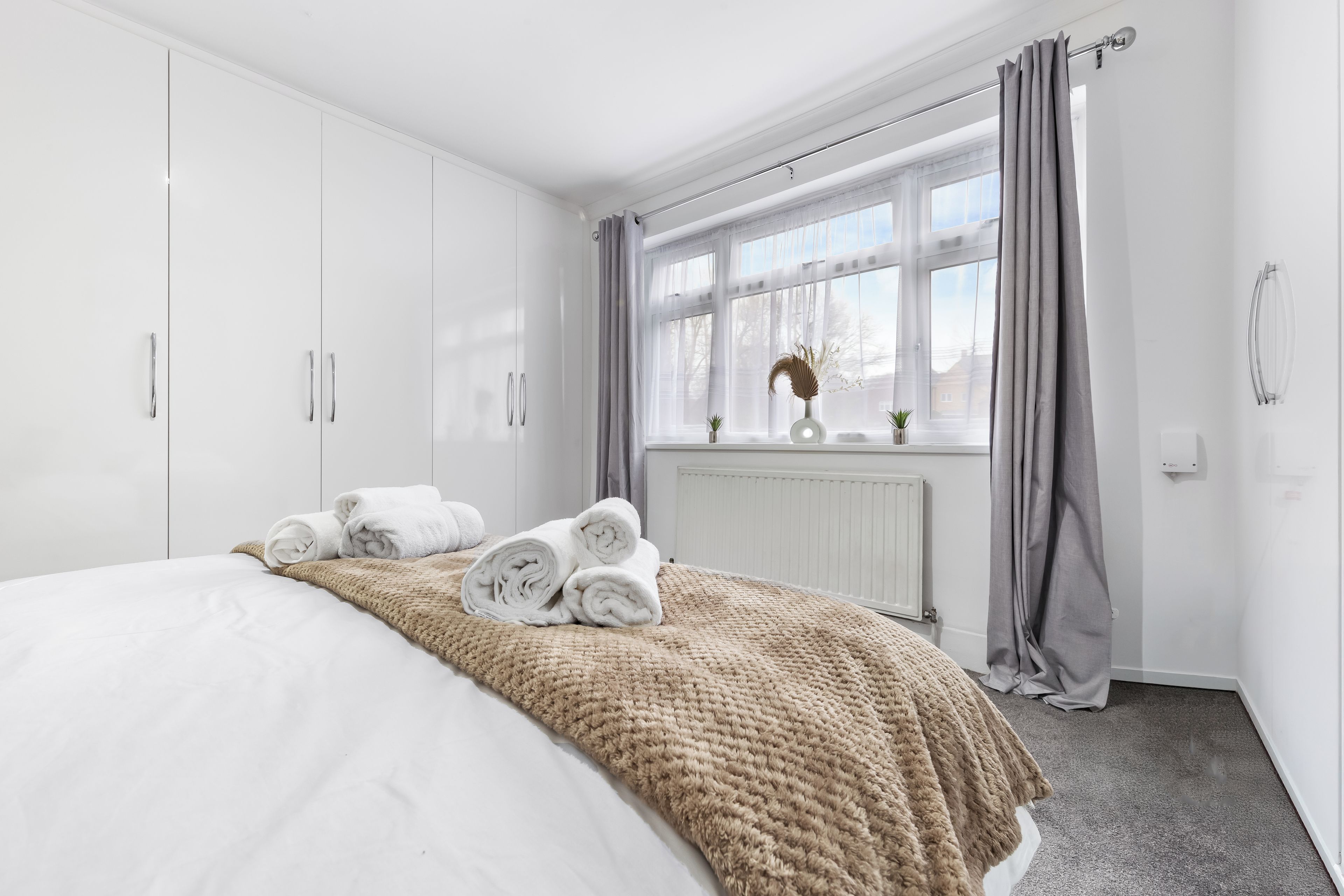 A neatly made bed with rolled towels, a window with curtains, and a radiator.