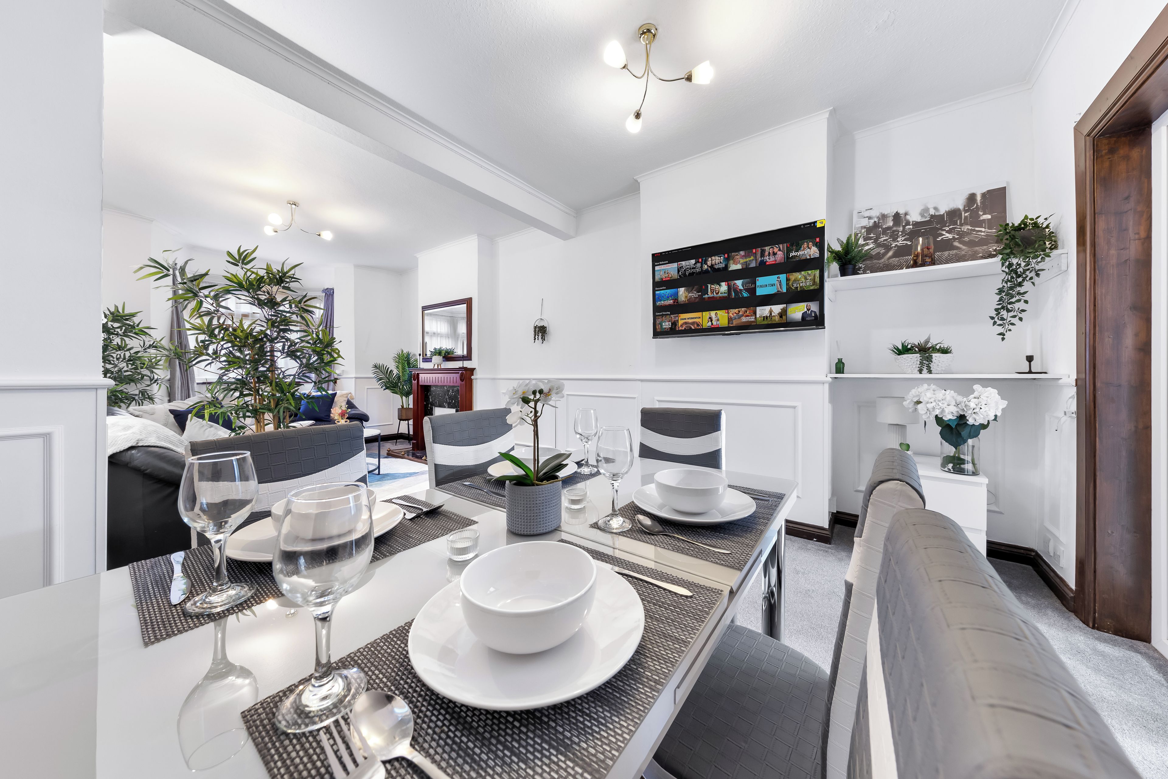 Un espace repas moderne avec une longue table dressée pour quatre personnes, entourée de chaises. On y trouve également un canapé et une petite table d’appoint sur laquelle est posé un vase de fleurs.
