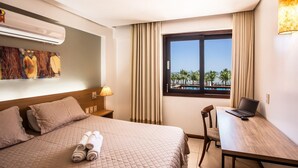 Habitación de hotel con cama, escritorio con una computadora portátil, ventana con vistas a palmeras y un equipo de aire acondicionado.