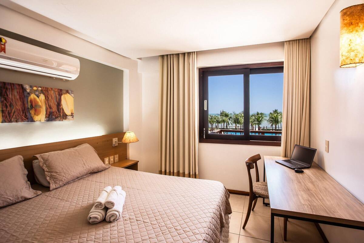 Ein Hotelzimmer mit Bett, Schreibtisch mit Laptop, Fenster mit Blick auf Palmen und Klimaanlage.