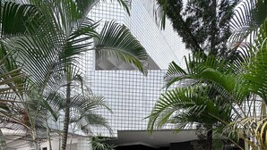 Uma passagem leva a um edifício com fachada moderna em estilo de grade, cercado por vegetação exuberante e palmeiras.