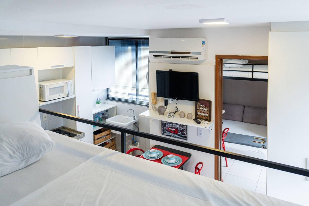 A compact hotel room with a bed, a small kitchenette with a microwave, a TV, and a window with blinds.