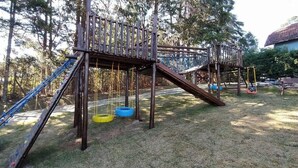 A wooden playground structure with slides and swings, surrounded by trees and grass.