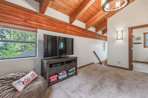 Un salón amplio con un televisor de pantalla plana grande, vigas de madera en el techo, una ventana con vistas a árboles y una zona de estar acogedora con una manta.