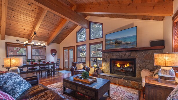 A spacious living room with a fireplace, wooden ceiling beams, large windows, and a stone fireplace surround.