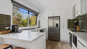 A modern kitchen with white cabinetry, a stainless steel refrigerator, a built-in oven, and a large window offering a view of trees. - Beachside Family Haven (Inverloch)