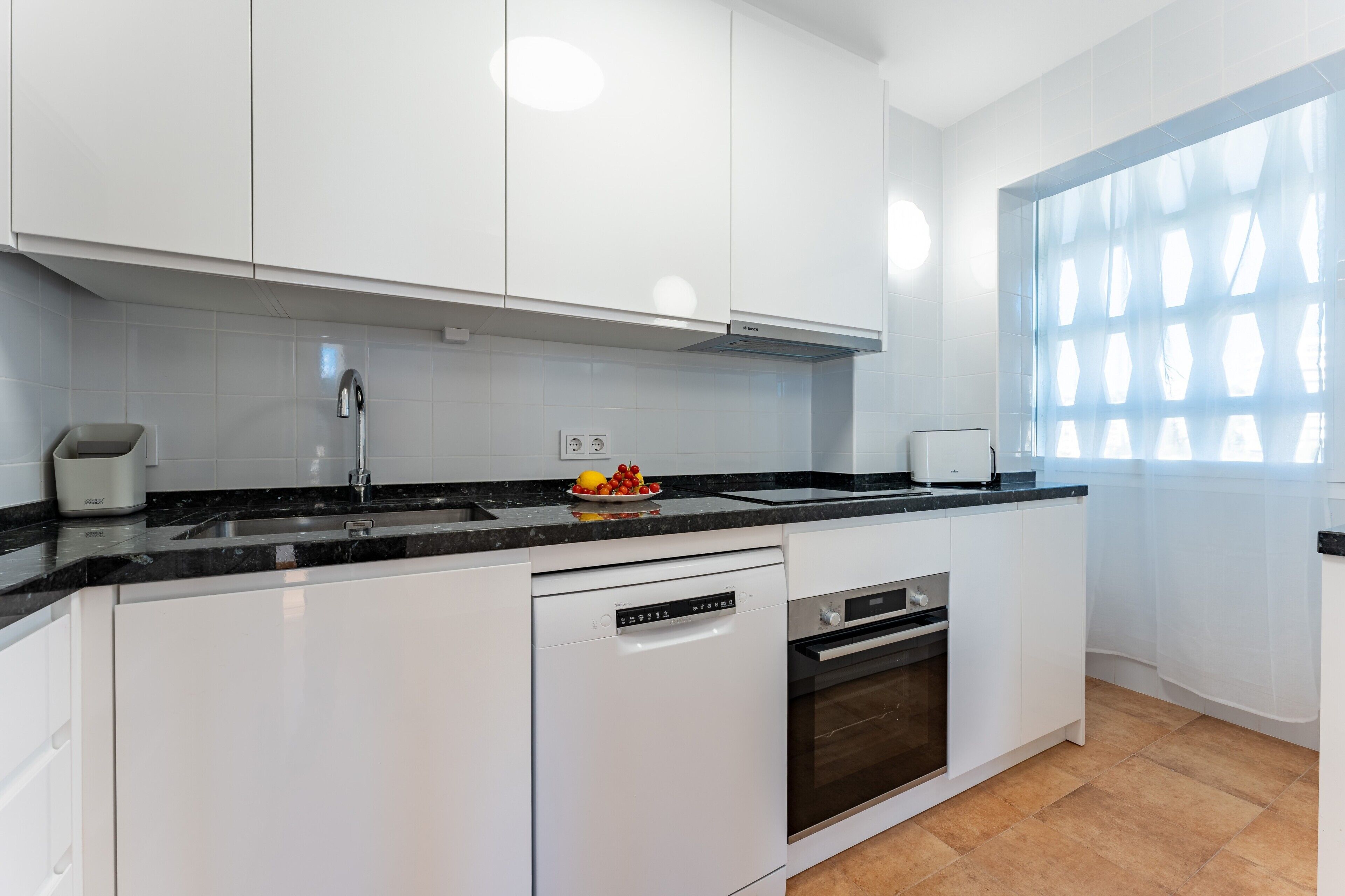 Une cuisine moderne avec des meubles blancs, un plan de travail noir, un lave-vaisselle, un four et une fenêtre ornée d’un motif géométrique.