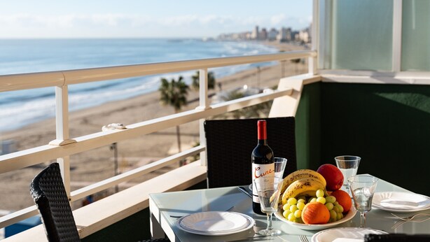 Una mesa con una botella de vino, una copa y un plato de fruta, con vista a una playa y una ciudad al fondo.