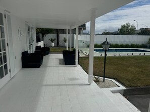 Ein überdachter Patio-Bereich mit weißem Bodenbelag, schwarzer Gartenmöbel und einem Pool im Hintergrund.