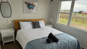 A bedroom with a bed, two bedside tables, a window with a view of vineyards, and a butterfly artwork on the wall. - Three Fifty Cottage (Havelock North)