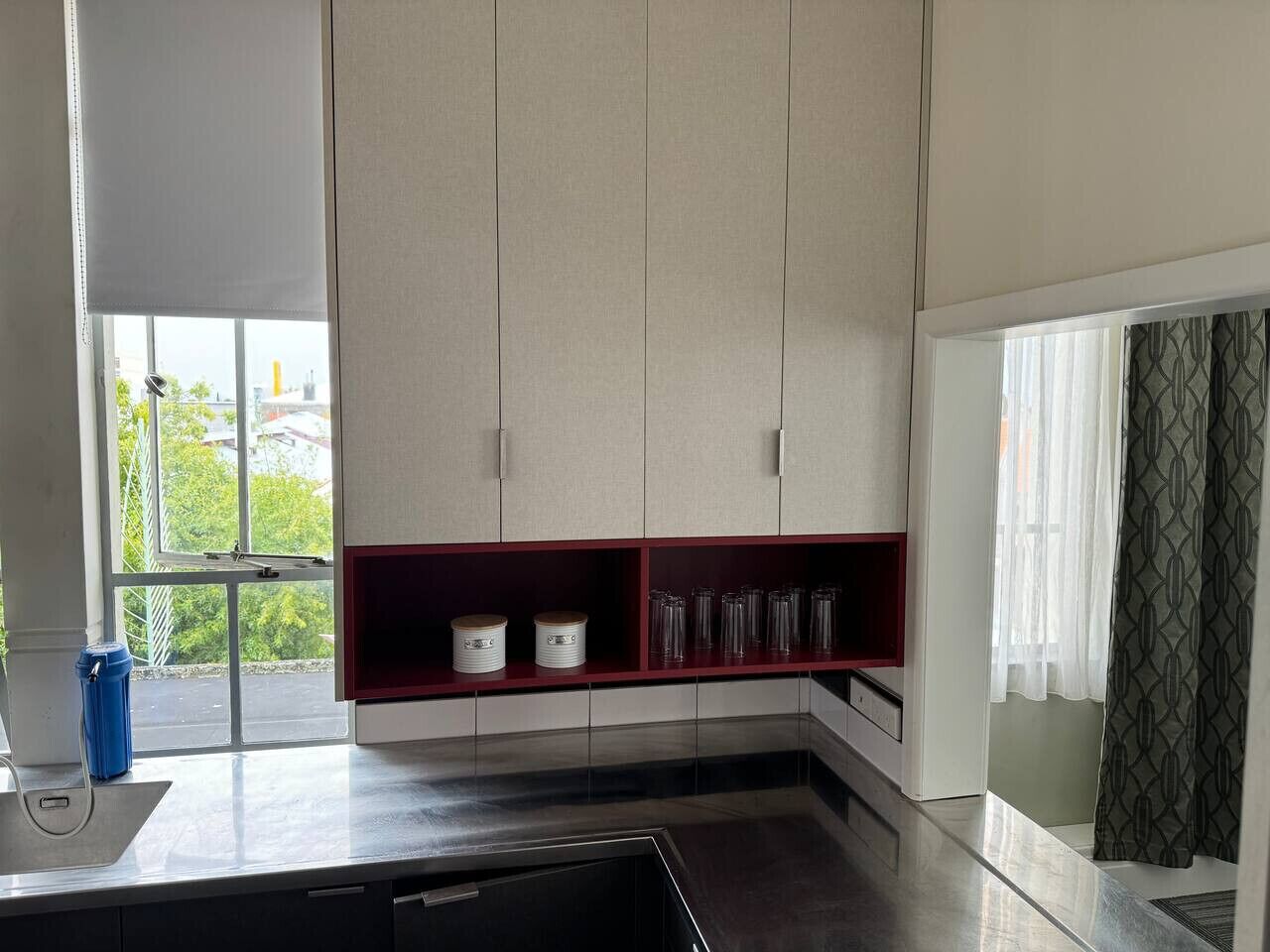 Een moderne keuken met een ingebouwde oven, een raam met uitzicht op groen en een rode plank met keukengerei.