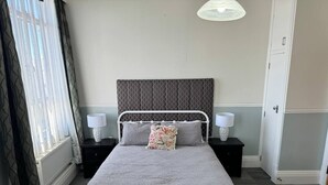 Ein ordentlich eingerichtetes Schlafzimmer mit einem Metallbettgestell, einer grauen Bettdecke, zwei Nachttischen mit Lampen, einem gemusterten Kissen und einem Fenster mit durchsichtigen Vorhängen.
