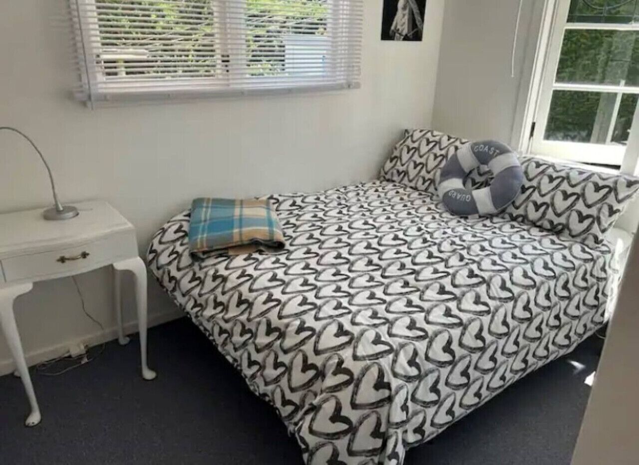 A bedroom with a bed featuring a heart-patterned bedspread, a bedside table with a lamp, and a window with blinds.