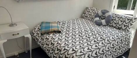 A bedroom with a bed featuring a heart-patterned bedspread, a bedside table with a lamp, and a window with blinds.