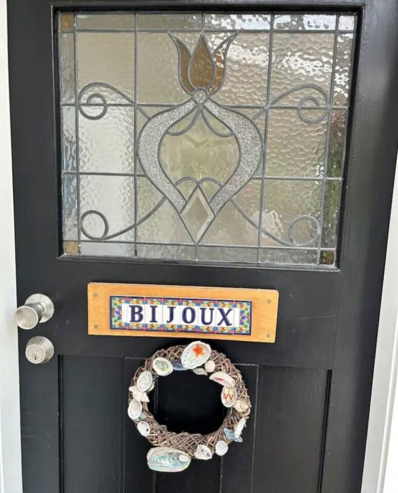 A black door with a glass panel featuring a decorative design, a sign reading "BIJOUX," and a wreath with various ornaments.