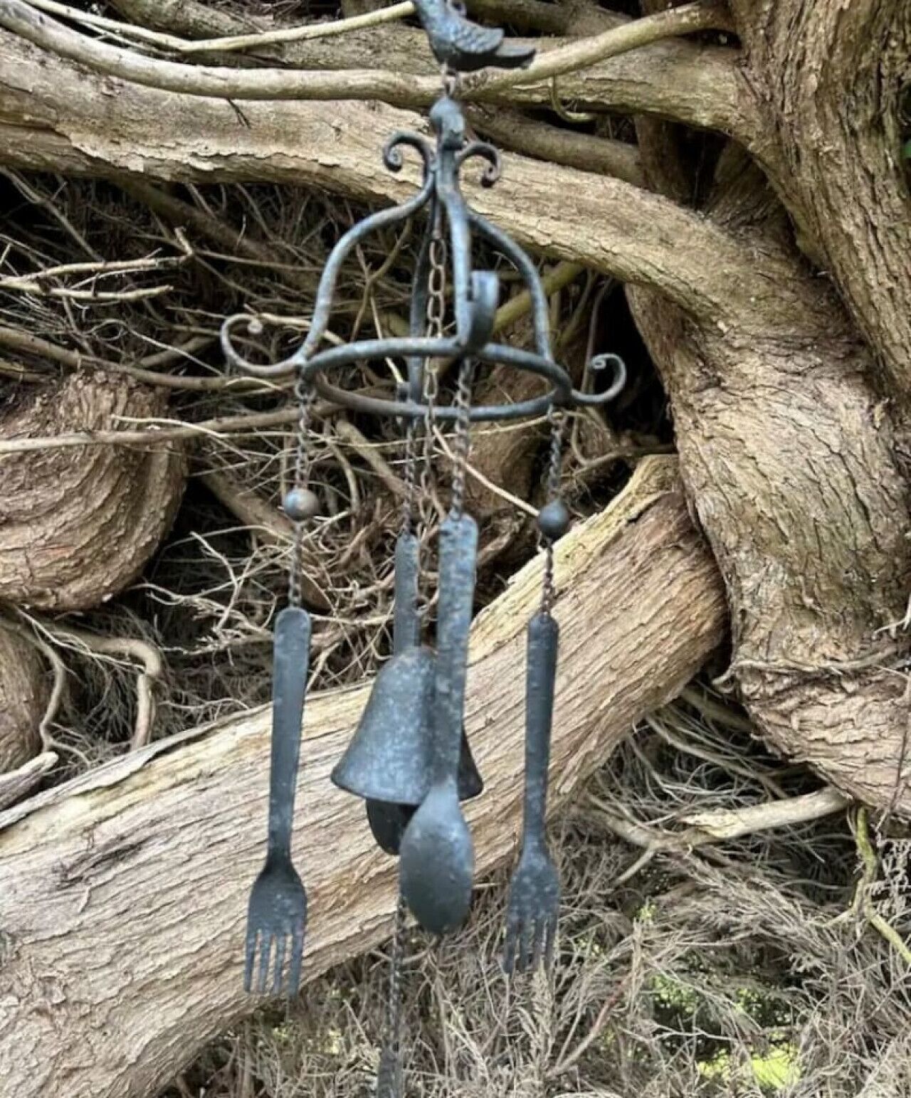A metal wind chime with a bird figure and three bells hanging from a chain, set against a backdrop of a large log and dry branches.