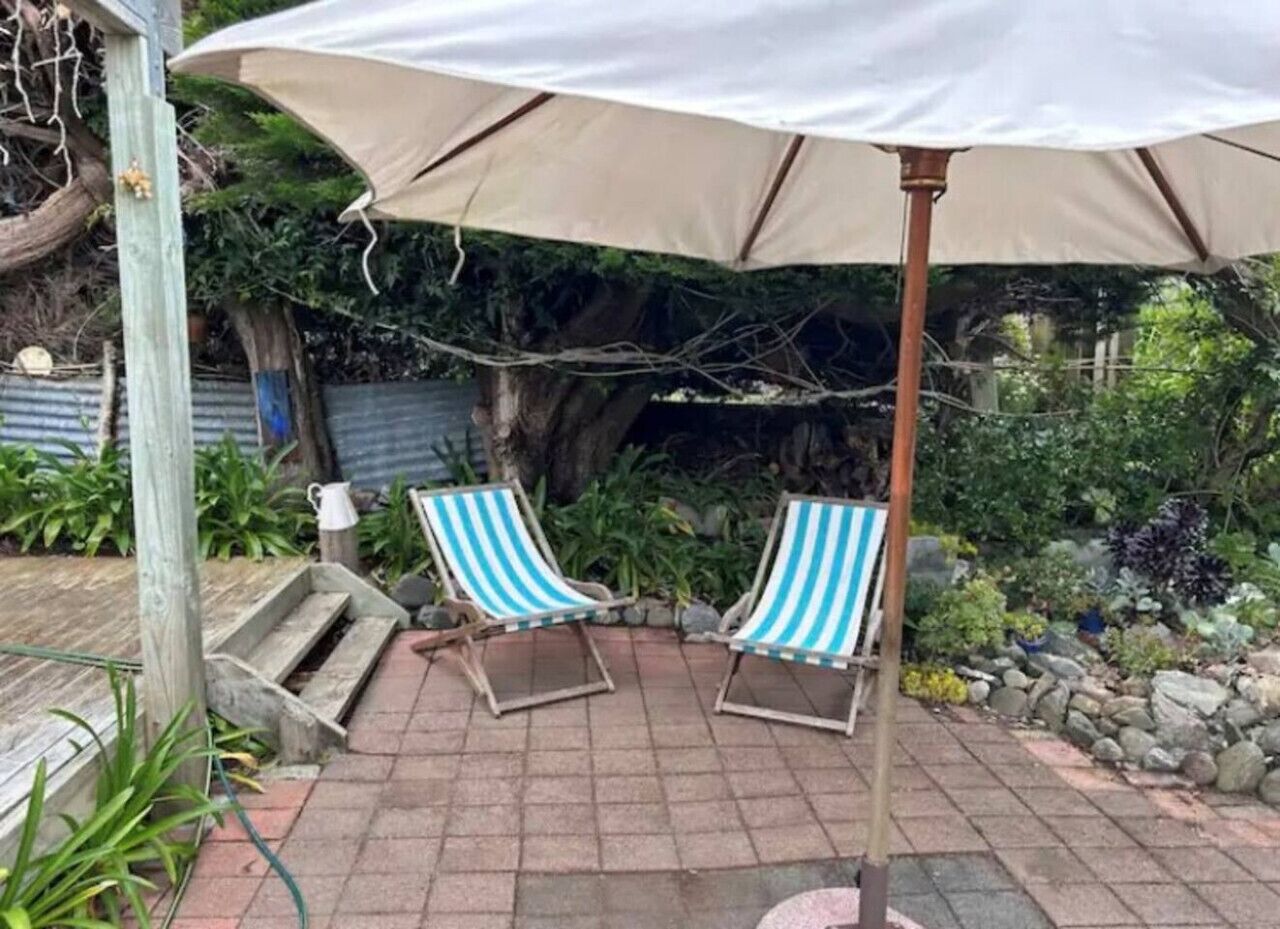 Una zona patio con due poltrone a righe sotto un ombrellone grande, circondata da un giardino con diverse piante e un muro in pietra.